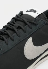 Sorte sneakers lavet af ruskind og syntetiske materialer, med sorte snørebånd, et hvidt Nike-logo på en sort tunge og en kontrasterende grå swoosh.