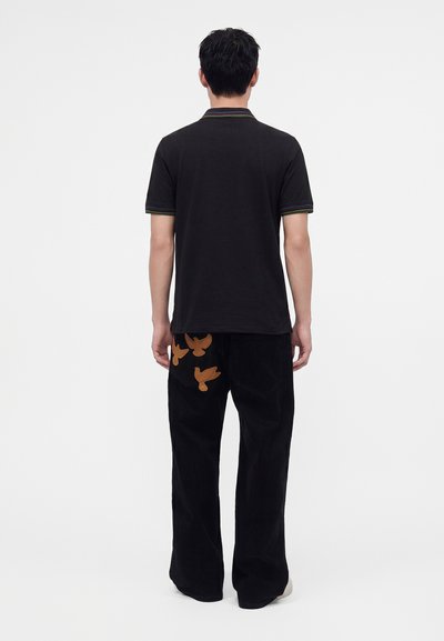 Homme de dos portant un polo noir à manches courtes et un pantalon noir avec trois motifs d'oiseaux bruns sur la poche arrière droite.