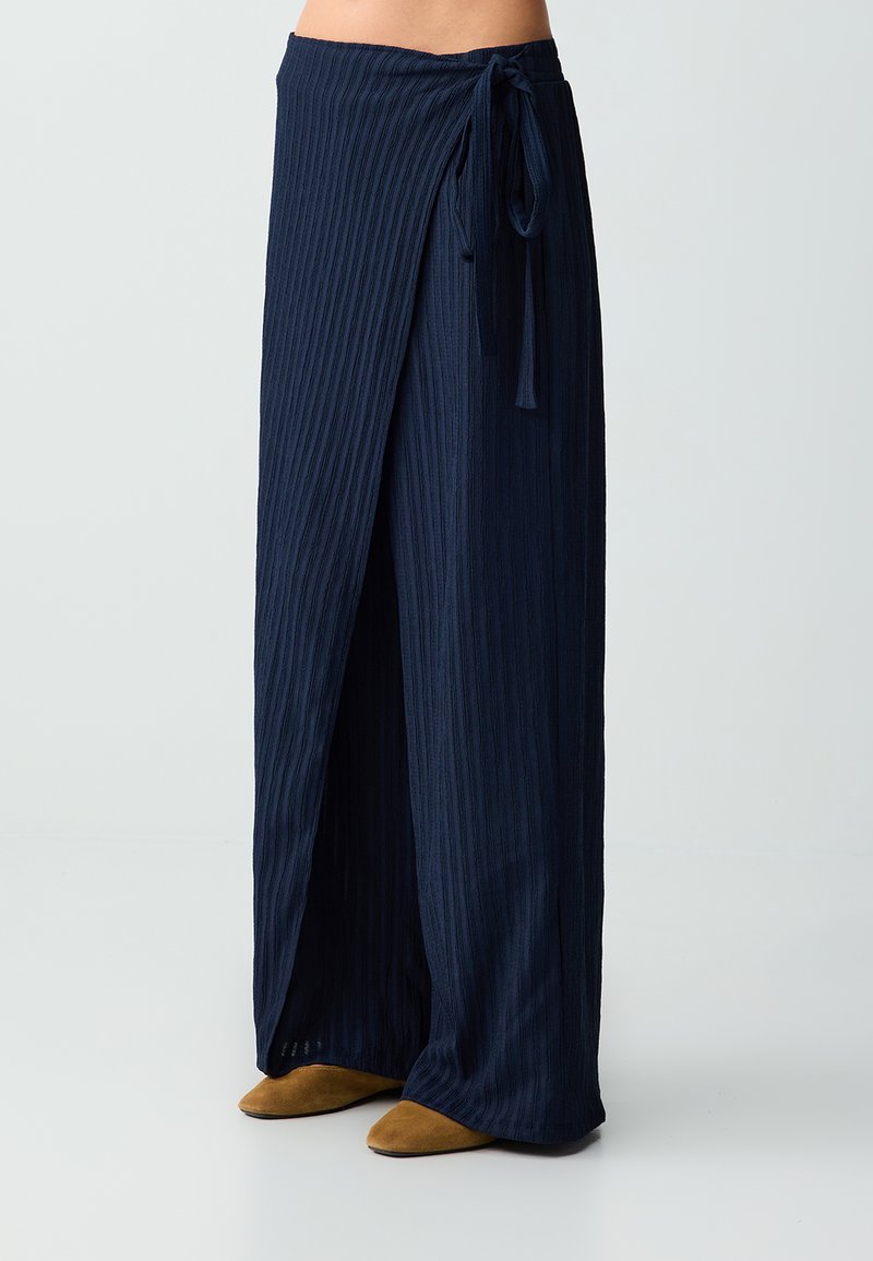 Jupe portefeuille côtelée bleu marine nouée à la taille, portée avec des chaussures pointues marron, montrée de la taille aux pieds sur un fond uni.