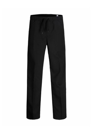 Garçon blond souriant debout, bras détendus, portant un polo vert olive à manches courtes, un pantalon noir et des chaussures beiges sur un fond blanc uni.