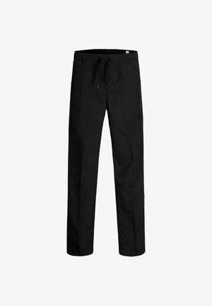 Garçon blond souriant debout, bras détendus, portant un polo vert olive à manches courtes, un pantalon noir et des chaussures beiges sur un fond blanc uni.