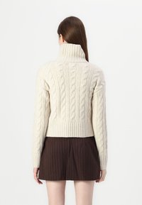 Suéter creme de malha com padrão de cabos, com um colarinho alto, bainha canelada e detalhes texturizados, emparelhado com uma saia castanha com riscas.