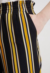 Svarta byxor med vertikala ränder i gult, vitt och marinblått. Elastisk midja och sidoficka synlig på höger sida.