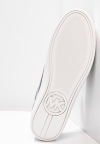 Sohle eines weißen Sneakers, schräg gegen eine weiße Wand positioniert, mit einer strukturierten Grifffläche und einem runden "MK"-Logo nahe der Ferse.