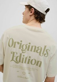 Kreminės spalvos trumpa rankovėmarškinė su dideliu žaliu tekstu "Originals Edition" ant nugaros, laisvo kirpimo ir minkštos audinio tekstūros.