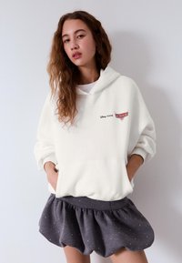 Fehér kapucnis pulóver kenguruzsebbel és "Disney Pixar Cars" logóval. Sötétszürke szoknyával párosítva, fodros szegéllyel és fehér pöttyökkel.