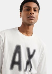 T-shirt bianco a maniche lunghe con grande grafica nera "AX" stampata in texture a pois. Colletto rotondo, materiale in morbido cotone.
