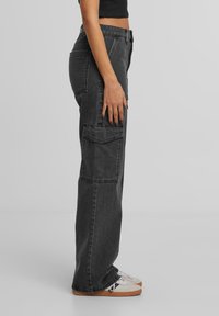 Pantalons cargo en denim noir avec une coupe décontractée, dotés de plusieurs poches et d'une texture légèrement décolorée. Portés avec des baskets de couleur claire.