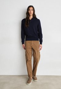 Ναυτικό πόλο πουλόβερ, καφέ cargo παντελόνι με πλαϊνές τσέπες, υφή υλικού, slip-on παπούτσια με καφετί λεπτομέρειες, όρθιος σε απλή επιφάνεια.