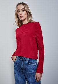 Rotes geripptes Langarmoberteil mit Rundhalsausschnitt und drei metallischen Knöpfen an der linken Schulter, kombiniert mit blauen Jeans.