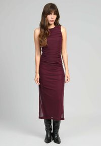 Femme avec de longs cheveux bruns portant une robe bordeaux sans manches et ajustée, ainsi que des bottes noires jusqu'aux genoux, se tenant devant un fond gris.