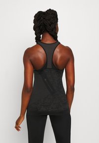 Mujer atlética con el pelo trenzado, vistiendo una camiseta de tirantes de malla negra y leggings negros, vista desde atrás.