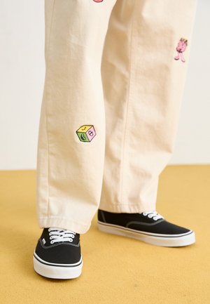 Pantalon en coton beige avec des motifs brodés ludiques, incluant un personnage rose et un bloc de couleur, associé à des baskets noires à lacets.