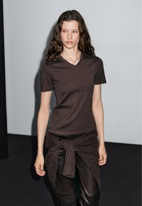 T-shirt marrone a scollo a V realizzato in tessuto morbido, abbinato a pantaloni scuri e strutturati e a un maglione annodato in vita. Design semplice, forma aderente.