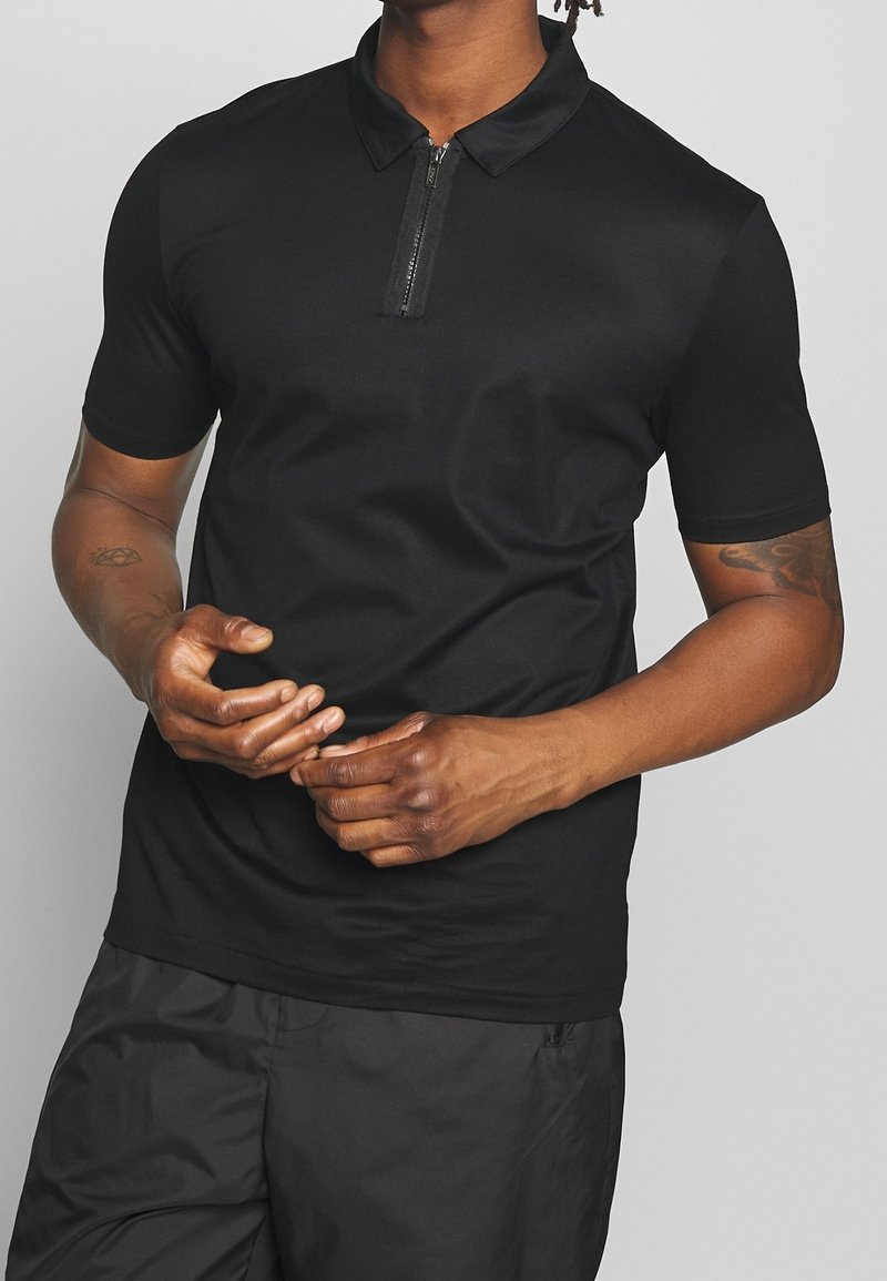 Homme portant un polo noir à manches courtes avec une fermeture éclair sur le devant et un pantalon noir, debout les mains jointes devant.