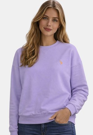 Junge Frau mit langen blonden Haaren, die einen hellvioletten Pullover und blaue Jeans trägt, lächelt mit einer Hand in der Tasche.