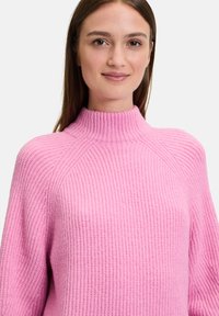 Roze gebreide trui met hoge hals, met een ribbeltextuur en een ontspannen pasvorm. Het ontwerp is eenvoudig met minimale details.