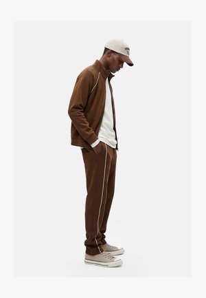 Man draagt een bruin trainingsjack en broek met witte zijstrepen, wit hemd, beige sneakers en een beige pet, staat met handen in de zakken.