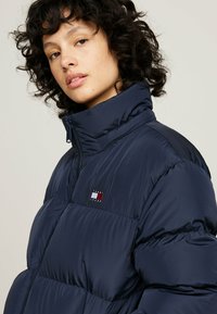 Marine pufferjack met een horizontaal gequilt ontwerp, hoge kraag en een merkembleem met rood, wit en blauwe accenten. Ritssluiting.