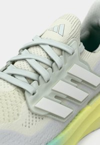 Chaussure de sport gris clair avec empeigne en mesh, lacets, et un design blanc à trois bandes. La semelle intermédiaire amortissante se transforme en un dégradé de jaune et vert.