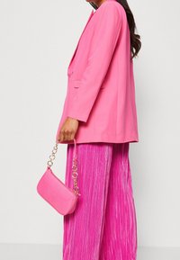 Jasně růžový blejzr s klopami, kombinovaný s plisovanými růžovými kalhotami. Drží malou, texturovanou růžovou kabelku s řetízkem.