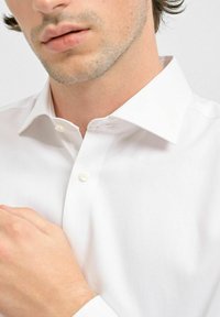 Hvid skjorte med klassisk krave, glat tekstur og to synlige knapper ved halsen. Modellens hånd hviler på skjortens stof.