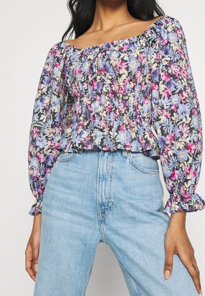 Žena nosící květovanou halenku s odhalenými rameny a nafouklými dlouhými rukávy, a světle modré džíny s vysokým pasem, zobrazená od ramen po boky.