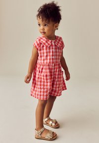 Bambina in piedi, indossa un vestito a quadretti rosso e bianco con tasche e sandali con fibbia dorata, guarda leggermente di lato.