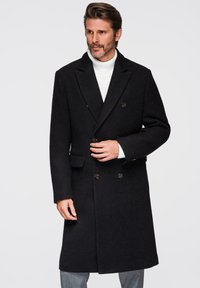 Abrigo de lana negro de doble botonadura con botones grandes, cuello con muesca y bolsillos laterales. Lleva puesto un suéter de cuello alto blanco.