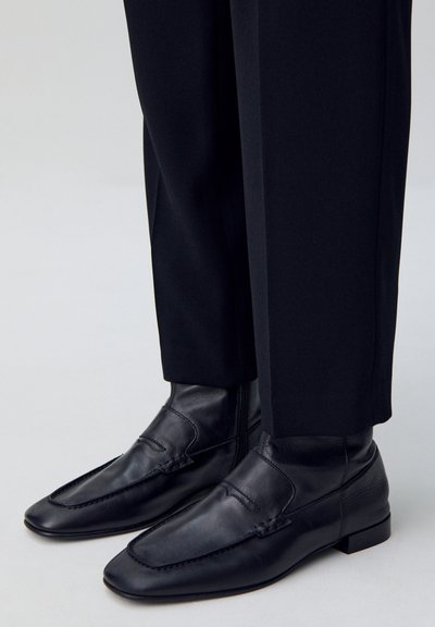 Mocasines de cuero negro con una textura suave, puntera afilada, costuras decorativas y un tacón bajo en bloque, combinados con pantalones negros.