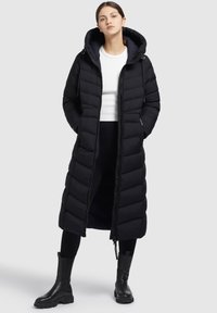 Palton puf negru cu glugă, având secțiuni matlasate, fermoar frontal complet și o siluetă lungă. Purtați peste un top alb și pantaloni negri.