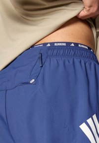 Tengerészkék futónadrág rugalmas derékpánttal, amelyen a "RUNNING" felirat található, oldalzsebbel és texturált anyagból. A nadrág felett egy világos színű alsóing látható.