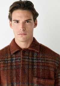 Brown plaid shirt jacket made of textured fabric, featuring a collared neckline, button closure, and a single chest pocket.