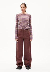 Frau in einem lila-beigen gestreiften Langarmpullover, weit geschnittenen bordeauxfarbenen Hosen und schwarzen Mokassins vor einem weißen Hintergrund.