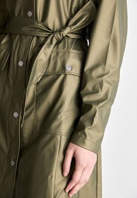 Olive green, waterproof jacket with a tie belt, bracelet sleeves, and metal snap buttons. Features a pocket on the front. Smooth texture.