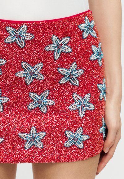 Κόκκινη μίνι φούστα διακοσμημένη με μπλε και λευκά πιασμένα στο χέρι μοτίβα σε σχήμα αστεριού με λουλούδια, φορεμένη από άτομο με λευκή μπλούζα.