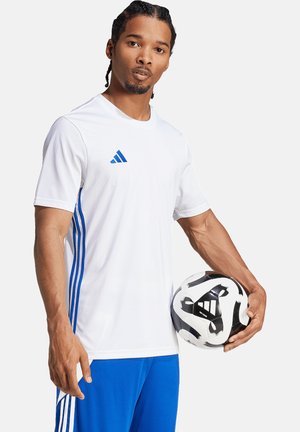 Weißes Sportshirt mit blauen Akzenten und drei Streifen, kombiniert mit blauen Shorts. Hält einen schwarz-weißen gemusterten Fußball.
