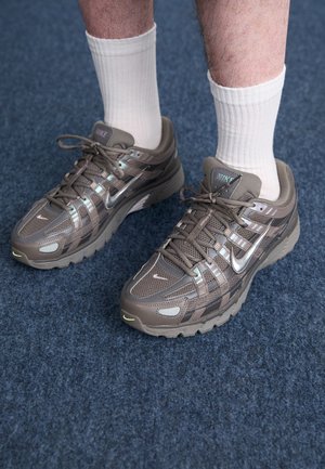 Personne portant des baskets Nike marron et des chaussettes blanches côtelées, debout sur un sol recouvert de moquette bleue.