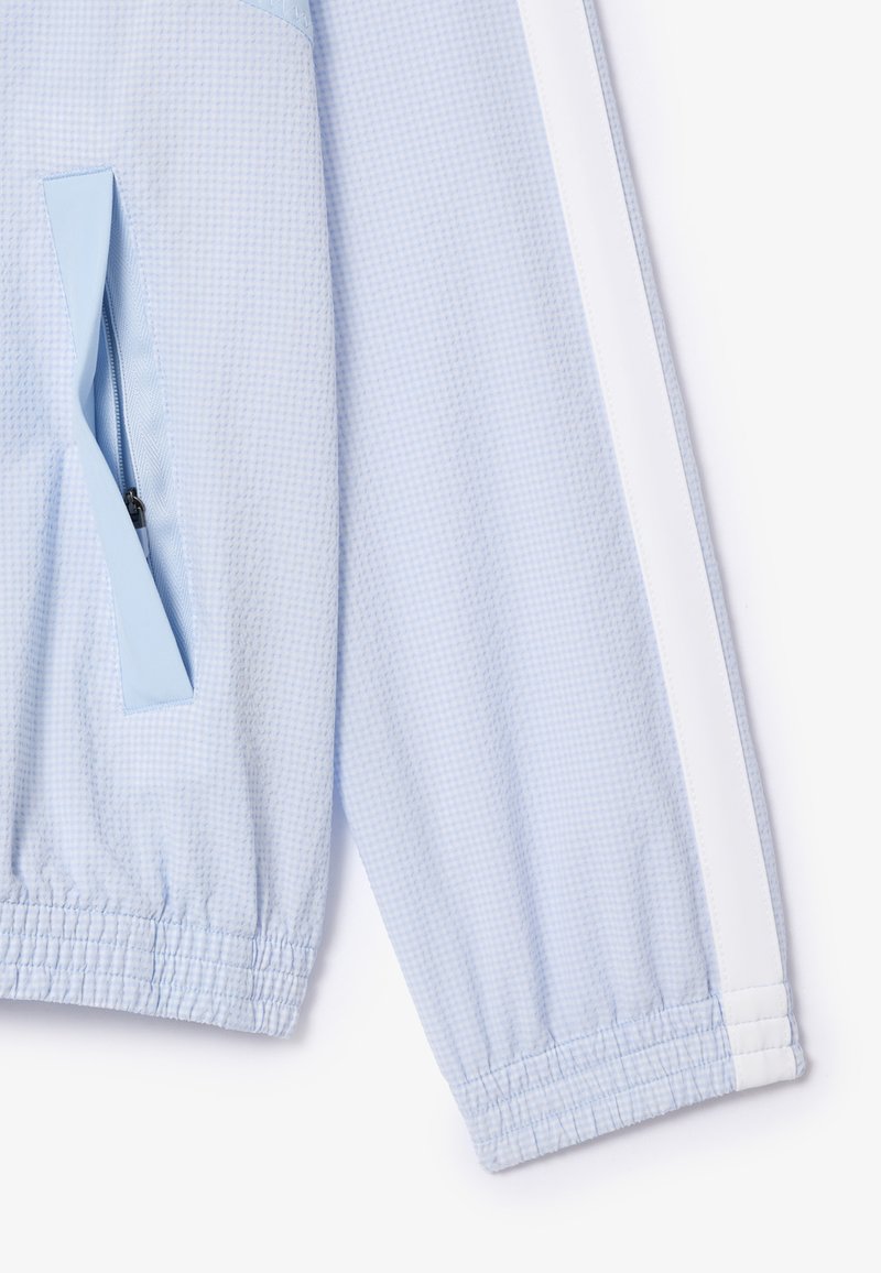Manche de veste texturée bleu clair avec poignet élastique, bande blanche le long du bras et poche zippée avec rabat en tissu.