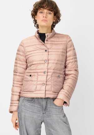 Frau trägt eine hellrosa gesteppte Jacke mit Druckknöpfen, schwarzer Innenschicht und verblassten grauen Jeans, steht mit einer Hand in der Hosentasche.