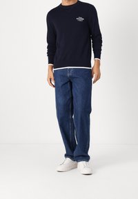 Sweat-shirt en coton bleu marine avec logo blanc sur la poitrine, associé à un jean en denim bleu coupe décontractée et des baskets blanches. Design simple et look décontracté.