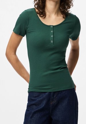 Vrouw draagt een aangesloten, geribd groen shirt met korte mouwen, voorzien van vier zilveren knopen, en donkerblauwe spijkerbroek, poseert met handen achter de rug.