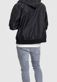 Svart nylon bomberjacka med huva, elastisk fåll och muddar, visad bakifrån. Tillsammans med ljusgrå tajta jeans.