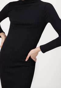 Abito nero a costine con maniche lunghe e collo alto, caratterizzato da una silhouette aderente e un delicato motivo testurizzato. Tessuto con finitura liscia.