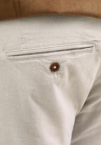 Poche en tissu gris clair d'un pantalon avec un détail de bouton marron, présentant un tissage texturé et des bords cousus.