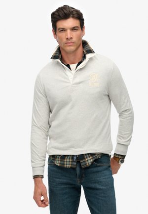Lichtgrijze longsleeve polo met een witte kraag en borstembleem. Daaronder steekt een zwart-geel geruit shirt uit. Gecombineerd met een blauwe spijkerbroek.