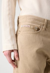 Pantaloni in denim beige con accessori in argento, una tasca frontale e un'etichetta rossa "Calvin Klein" sulla cintura. Indossati con un top di colore chiaro.