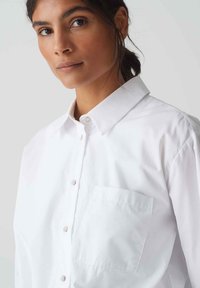 White button-up shirt with a pointed collar, long sleeves, single chest pocket, and a smooth cotton texture.