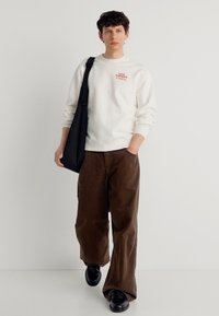 Νεαρός που περπατά φορώντας λευκό φούτερ, καφέ παντελόνι με φαρδιά πόδια, μαύρα παπούτσια και κρατώντας μια μαύρη τσάντα ώμου σε λευκό φόντο.