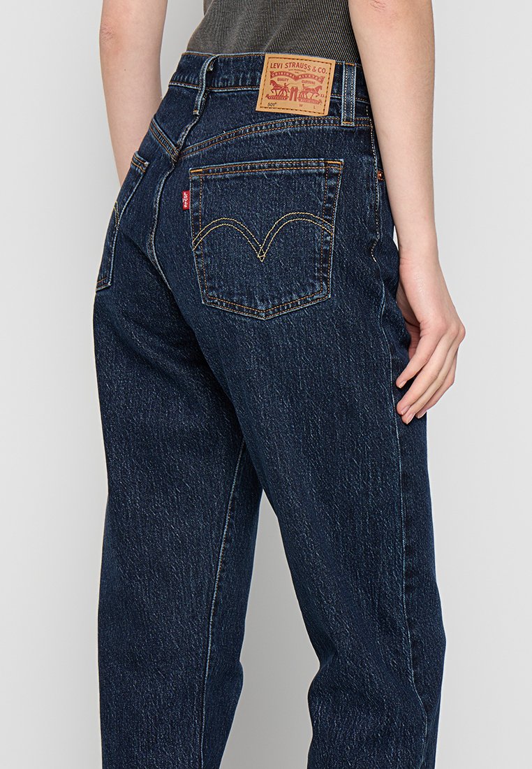 Jean en denim bleu foncé à taille haute, avec une étiquette en cuir beige à l'arrière, des coutures jaunes contrastantes et des poches arrière classiques.